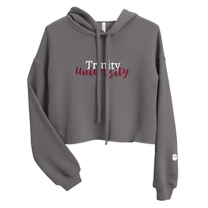 May include: A cropped, charcoal gray hoodie with a drawstring hood. The front displays the text "Trinity University" in white and maroon. A paw print logo is on the left sleeve.