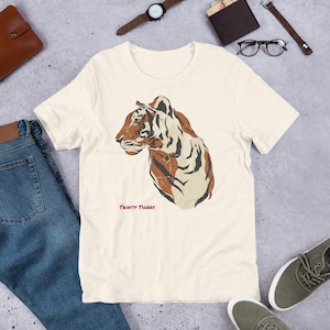 May include: Cream-colored t-shirt featuring a distressed tiger head graphic in shades of brown and cream. The text "Trinity Tigers" is printed in pink below the tiger. The shirt is displayed with blue jeans, a watch, and other accessories.