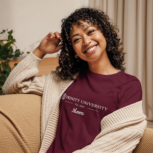 Può includere: Una maglietta a maniche lunghe bordeaux con la scritta bianca "Trinity University Est. 1869 Mom". La maglietta è indossata con un cardigan beige.