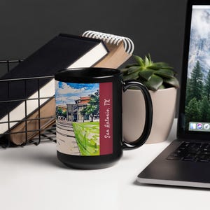May include: Black coffee mug with a colorful illustration of a building and the text "San Antonio, TX" in white. The mug is on a white surface with a laptop, a plant, and a notebook in the background.