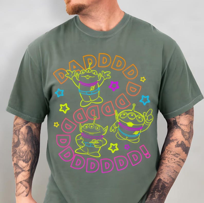 May include: Green t-shirt featuring three cartoon aliens with three eyes, outlined in neon green, blue, and pink. The word "DAD" is repeated around the aliens in a circle, with exclamation point. Colorful star shapes are also included.