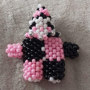 May include: A handmade beaded figure in a doll-like design. The figure is primarily pink, black, and white, with silver accents. The figure has a conical head and a body with arms and legs, all made from small beads.