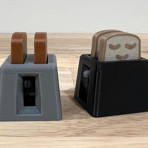 Op de afbeelding: Twee speelgoedtoasters, één grijs en één zwart, elk met sneetjes toast. De grijze broodrooster heeft twee bruine toastjes. De zwarte broodrooster heeft drie sneetjes toast met lachende gezichten.