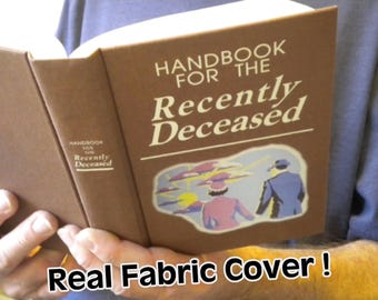 May include: A brown book with the title "Handbook for the Recently Deceased" on the cover. The cover has a fabric texture and features a colorful illustration of two people in a landscape.