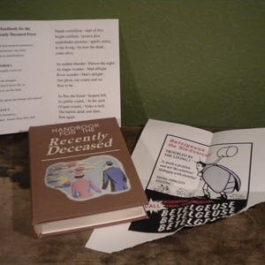 May include: A brown book titled "Handbook for the Recently Deceased" with a black and white illustration of a woman and a man on the cover. The book is sitting on a wooden surface with a folded white paper and a white paper with black text that reads "Betelgeuse the Bio-Exorcist" and a black and white illustration of a beetle.