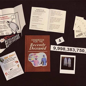 May include: A red book titled "Handbook for the Recently Deceased" with a sticker of a couple in hats on the cover. There are other items on the table, including a folded paper with the text "Betelgeuse" and a Polaroid photo of two white ghosts.