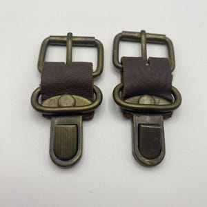 Puede incluir: Dos conectores de hardware a juego con correas de cuero marrón oscuro aseguradas por hebillas de metal en tono latón antiguo y clips de resorte rectangulares con una textura acanalada vertical en la parte frontal.