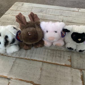 Puede incluir: Un conjunto de cuatro juguetes de peluche que incluye un perro blanco y negro, un alce marrón con astas, un cerdo rosa claro y una oveja blanca con cara negra. Cada pequeño animal de peluche tiene una etiqueta de la marca Puffling al costado.
