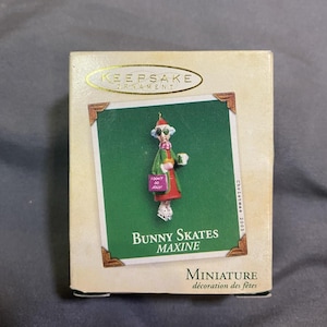Può includere: Scatola di ornamenti natalizi Keepsake con una miniatura di un coniglietto sui pattini. Il coniglietto indossa un cappotto verde, una gonna rossa e tiene in mano un cartello con la scritta "I DON'T DO JOLLY". La scatola recita "Bunny Skates Maxine".