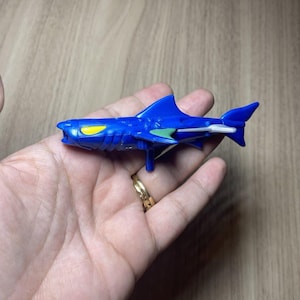May include: A small blue plastic shark toy featuring a yellow eye, a green side fin, and a silver tail fin. The shark has a moulded body with a small peg extending from the underside, designed for play or display as a collectible action figure.