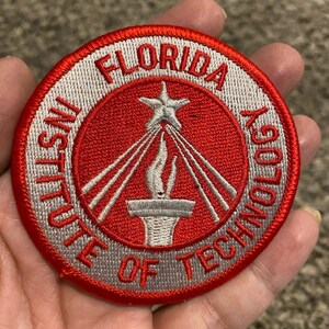 Könnte beinhalten: Ein runder, gestickter Aufnäher mit der Aufschrift "FLORIDA INSTITUTE OF TECHNOLOGY". Der Aufnäher hat einen roten Hintergrund mit einem weißen und silbernen Design aus Stern, Fackel und Strahlen. Der Rand ist rot und silber.