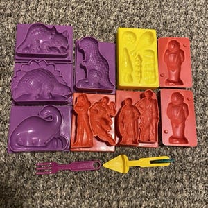 May include: A collection of plastic moulds in purple, yellow, and red, featuring various dinosaur shapes, human figures, and small food items. Included are a purple fork and a yellow spatula tool, all designed for shaping clay or dough materials.