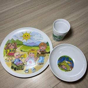 May include: A white plastic dinnerware set consisting of a plate, a bowl, and a cup. The plate features a colorful illustration of four children playing outdoors under a yellow sun. The bowl has a small circular graphic of a child in a tent.