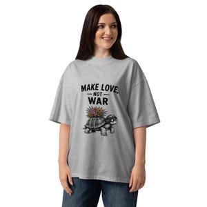 May include: A heather gray short-sleeve t-shirt featuring a black graphic of a turtle wearing a helmet with a bouquet of colorful flowers on its shell. Above the turtle, the text reads "MAKE LOVE, NOT WAR" in bold black capital letters.