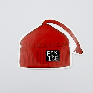 May include: A red beanie sticker with the text "FCK ICE" in a black rectangle. The beanie has a ribbed texture and a long, dangling cord. The sticker is on a white background.
