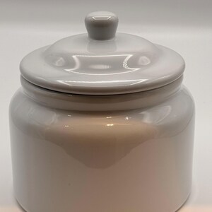 May include: A glossy white ceramic canister with a smooth, cylindrical body and a matching lid. The lid features a small, rounded knob handle at the centre. The entire piece has a reflective, uniform finish with no patterns or additional decorations.