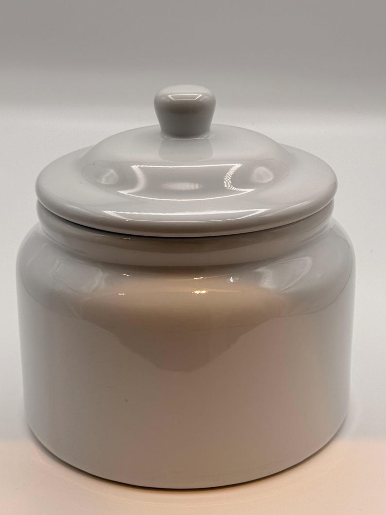 May include: A glossy white ceramic canister with a smooth, cylindrical body and a matching lid. The lid features a small, rounded knob handle at the center. The entire piece has a reflective, uniform finish with no patterns or additional decorations.