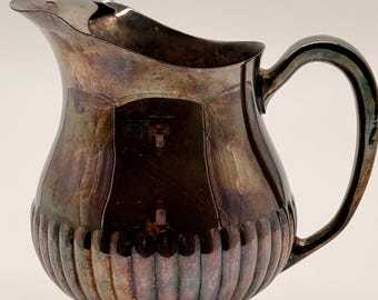 May include: A silver-plated pitcher with a tarnished, dark patina. The vessel features a wide, curved spout, a single arched handle, and a rounded body with vertical fluted detailing at the base, resting on a circular pedestal foot.