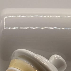 Puede incluir: Una mantequera de cer&aacute;mica blanca con tapa y asa. La tapa tiene forma curva y un asa peque&ntilde;a. El plato tiene forma rectangular. El interior del plato es de color amarillo claro.