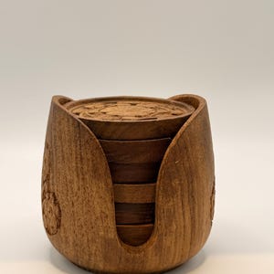 May include: A set of six wooden coasters and a holder. The coasters and holder are made of dark brown wood. The coasters have a carved design on the top. The holder has a carved design on the sides.