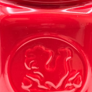 Könnte beinhalten: Nahaufnahme eines glänzend roten Keramiktopfes mit Deckel. Die Dose hat ein erhabenes Emblem auf der Vorderseite. Das Design ist ein stilisiertes Bild eines Pferdes. Die Dose hat eine glatte, reflektierende Oberfläche.