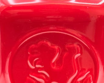 May include: Close-up of a glossy red ceramic jar with a lid. The jar features a raised emblem on the front. The design is a stylized image of a horse. The jar has a smooth, reflective surface.