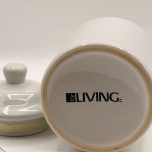 Puede incluir: Tarro de cer&aacute;mica blanca con tapa, con un pomo gris claro y un acento amarillo en el borde de la tapa. La base del tarro muestra el texto "for LIVING" en negro. El tarro est&aacute; sobre una superficie blanca.