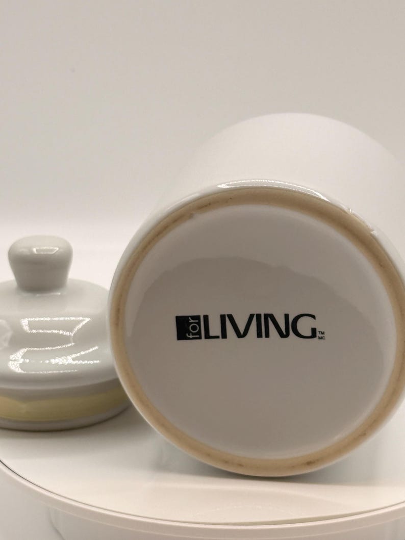 Puede incluir: Tarro de cer&aacute;mica blanca con tapa, con un pomo gris claro y un acento amarillo en el borde de la tapa. La base del tarro muestra el texto "for LIVING" en negro. El tarro est&aacute; sobre una superficie blanca.