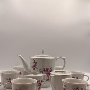 May include: A white porcelain tea set featuring a floral pattern. The set comprises a teapot, milk jug, sugar bowl, and cups with saucers. Each item is adorned with pink flowers and gold edging, offering a refined and traditional aesthetic.