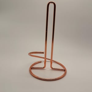 Puede incluir: Un soporte para papel de cocina de metal color cobre con una base circular y una varilla vertical alta en forma de U. La estructura está hecha de alambre fino, liso y pulido, creando un diseño simple y abierto que se mantiene en pie.