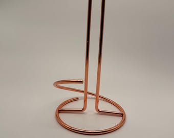 May include: A copper-colored metal paper towel holder featuring a circular base and a tall, U-shaped vertical rod. The structure is made of thin, smooth, polished wire, creating a simple, open design that stands upright on a flat surface.