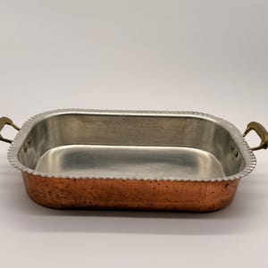 Puede incluir: Una bandeja de horno rectangular con exterior de cobre martillado e interior de metal plateado liso. La bandeja presenta un borde festoneado y dos asas de metal color latón fijadas a los lados con remaches visibles.