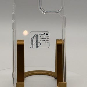 Op de afbeelding: Een doorzichtige, transparante beschermhoes voor een telefoon met een vierkante uitsparing voor de camera en een centrale sticker met een waarschuwingsicoon en de tekst: Note: Please peel off protective film before use, inclusief een illustratie.