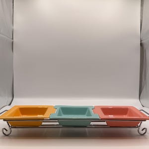 May include: A serving dish with three square ceramic bowls in yellow, teal, and coral, set on a chrome metal rack. The bowls are evenly spaced and have a slightly raised rim. The rack has decorative scrollwork on the ends.