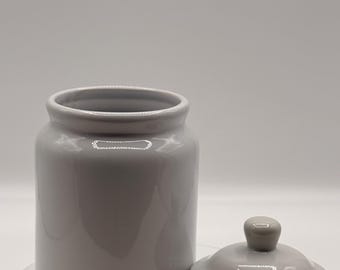 May include: A gray ceramic jar with a matching lid. The jar is cylindrical with a wide opening and a smooth, glossy finish. The lid has a small knob and a yellow rim. The jar and lid are on a white surface.
