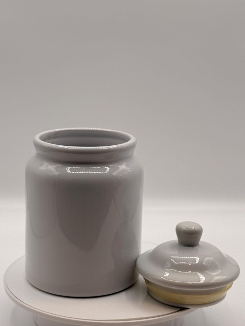 Puede incluir: Un tarro de cer&aacute;mica gris con tapa a juego. El tarro es cil&iacute;ndrico con una abertura ancha y un acabado liso y brillante. La tapa tiene un peque&ntilde;o pomo y un borde amarillo. El tarro y la tapa est&aacute;n sobre una superficie blanca.