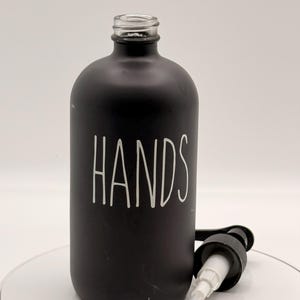 Puede incluir: Una botella de vidrio negro mate con la palabra "HANDS" en escritura blanca. Tiene un dispensador de bomba negro. La botella está diseñada para jabón líquido o loción.