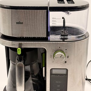 Puede incluir: Una cafetera Braun plateada y negra con jarra de cristal. El aparato cuenta con un depósito de agua transparente con marcas de hasta 50 oz, un dial circular para seleccionar el número de tazas y un panel de control digital con varios botones.