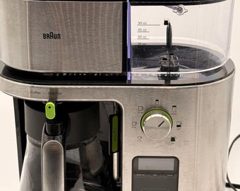 May include: A silver and black Braun coffee maker with a glass carafe. The machine features a clear water reservoir with markings up to 50 oz, a circular dial for cup selection, and a digital control panel with buttons for Over Ice, Light, Gold, and Bold.