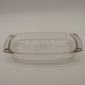 May include: A clear rectangular glass baking dish with rounded corners and integrated handles on both ends. The dish features a smooth, transparent surface and a shallow basin, designed for oven use with a sturdy rim and a flat bottom base.