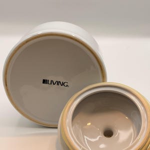 May include: A white ceramic canister with a matching lid, shown from an angle that reveals the underside of the lid and the base. The base features the text "for LIVING" printed in black. The ceramic has a smooth, glossy finish throughout.