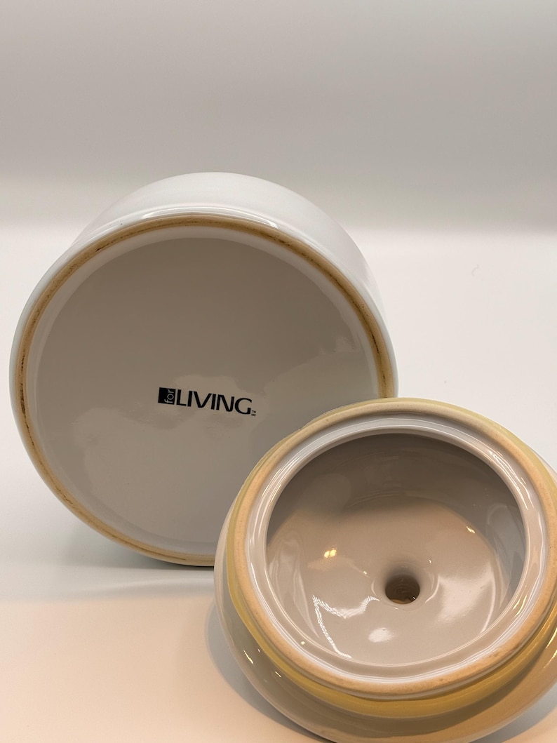May include: A white ceramic canister with a matching lid, shown from an angle that reveals the underside of the lid and the base. The base features the text "for LIVING" printed in black. The ceramic has a smooth, glossy finish throughout.