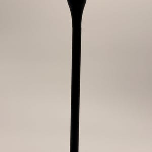 Puede incluir: Un candelabro de metal negro mate con un vástago cilíndrico vertical y delgado que se eleva desde una base circular, rematado con un receptáculo acampanado en forma de copa diseñado para sostener una vela cónica en posición vertical.