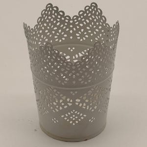 May include: A white cylindrical metal container featuring an intricate laser-cut lace pattern throughout the upper section and body. The top edge has a scalloped design, and the base is solid, providing a stable foundation for this open-top vessel.