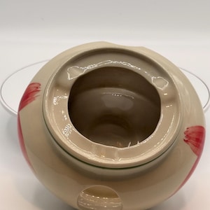 May include: A beige ceramic cookie jar with a lid, featuring red floral accents. The jar has a round shape with a recessed lid and a green trim. The interior is a darker beige colour.