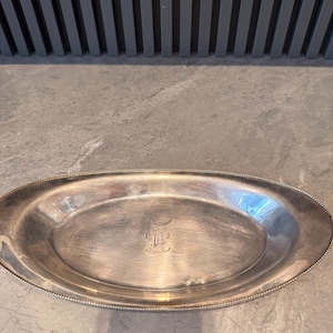 Peut inclure: Plateau de service ovale en argent avec un bord perlé et un monogramme au centre. Le plateau a une finition polie et reflète la lumière. Le plateau est probablement en argent ou en métal argenté.