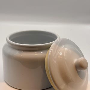 May include: A cylindrical ceramic canister with a glossy light beige finish and a matching removable lid. The lid features a small, rounded knob handle at the center. The interior of the canister is white, and the exterior has a smooth, reflective surface.