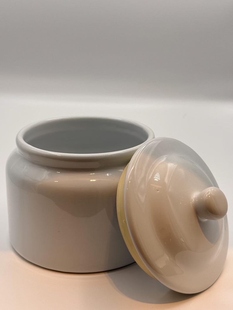 May include: A cylindrical ceramic canister with a glossy light beige finish and a matching removable lid. The lid features a small, rounded knob handle at the center. The interior of the canister is white, and the exterior has a smooth, reflective surface.