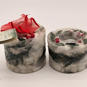 Peut inclure: Deux bougeoirs cylindriques en cire avec des aiguilles de pin vertes, des pierres blanches et des baies rouges incrustées. Le plus grand porte un ruban rouge et une étiquette Winter's Garden. Les deux ont un insert en verre pour bougie.