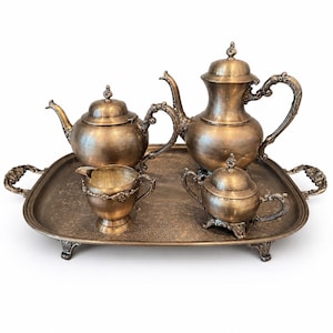 May include: A four-piece tea set in a tarnished brass finish, including a teapot, a coffee pot, a creamer, and a lidded sugar bowl. The pieces are arranged on a rectangular serving tray with ornate handles and an intricate embossed floral pattern.